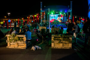 Zdjęcie z festiwalu, uczestnicy festiwalu w strefie chill out nocą, w tle podświetlona scena festiwalowa
