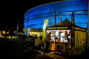 Zdjęcie z festiwalu, stoisko browaru bytów nocą, w tle podświetlona na niebiesko czasza stadionu