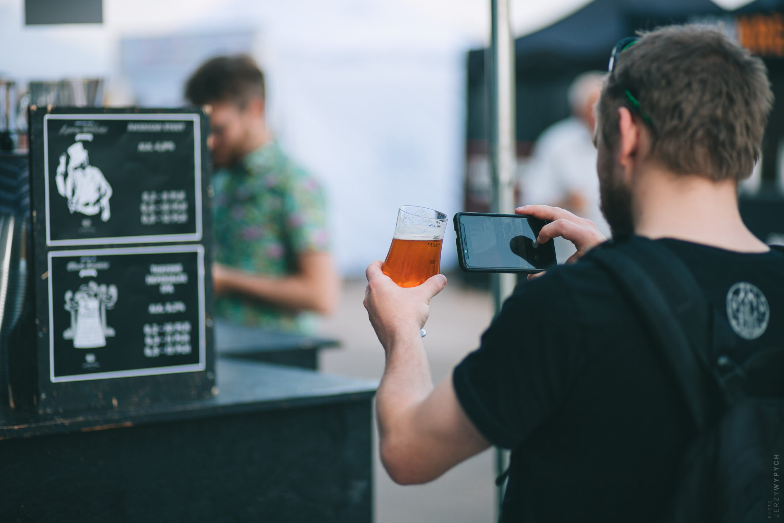 Zdjęcie z festiwalu, na zdjęciu mężczyzna robi zdjęcie szklance z piwem, w tle stoisko piwne